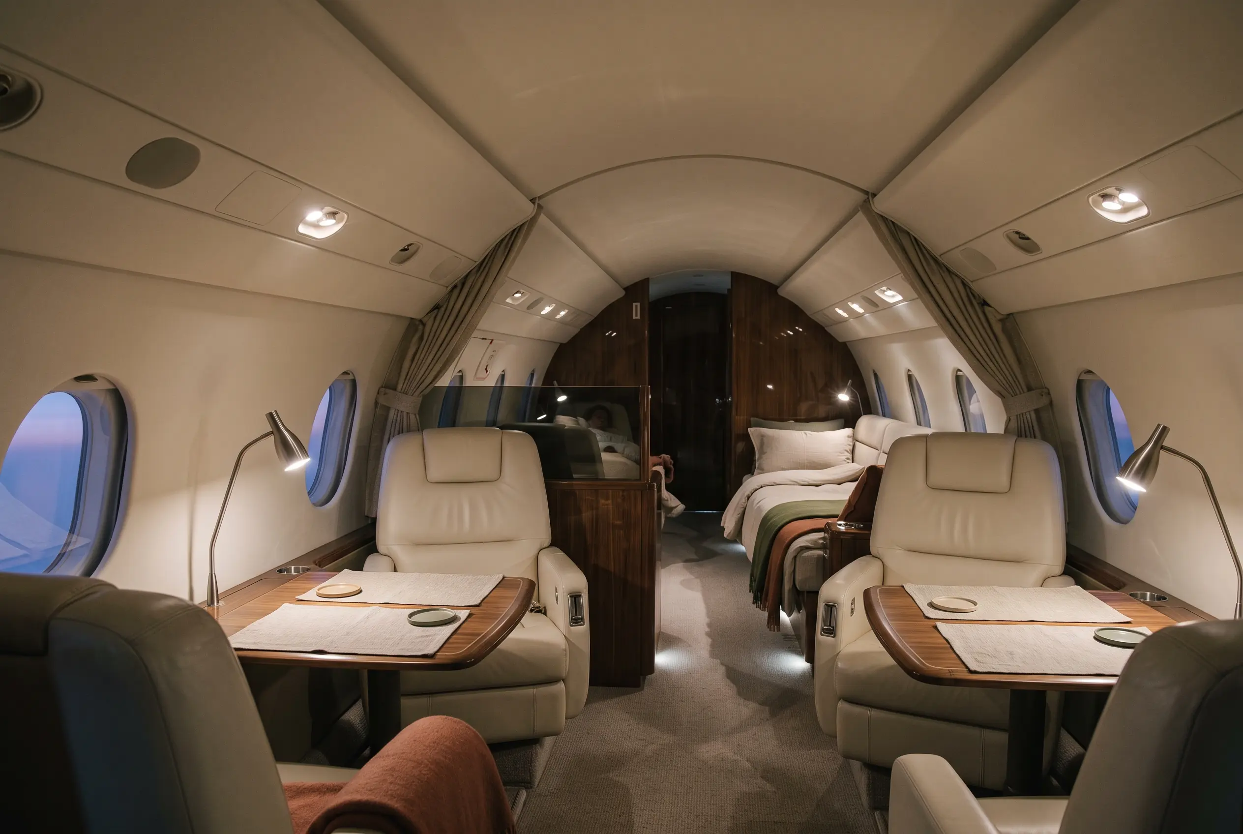 Long three-zone jet cabin at dusk, reading lamps glowing over club chairs and a made-up divan aft
